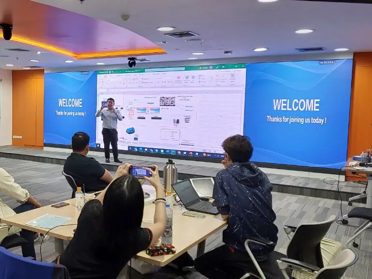 ผู้บรรยายยืนหน้าจอผนังขนาดกว้างที่ขึ้นคำว่า Welcome และผู้เข้าร่วมนั่งที่โต๊ะ