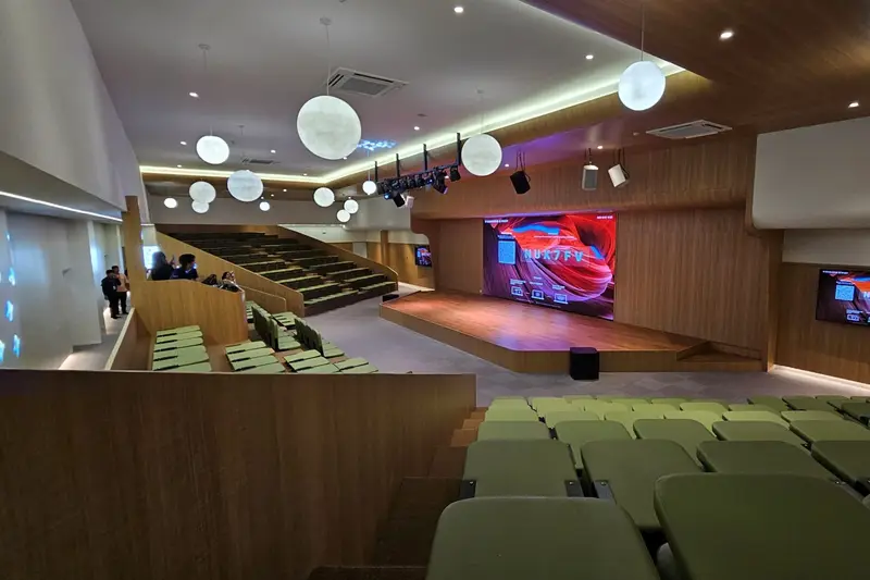 หอประชุมโรงเรียนที่มีที่นั่งสีเขียวแบบขั้นบันได หันหน้าไปทางจอ LED บนเวทีไม้ พร้อมลำโพงแขวนและไฟเวที