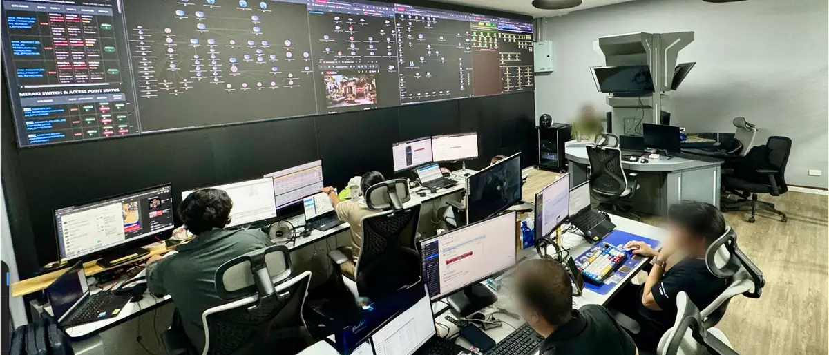 ทีมงานที่โต๊ะทำงานในห้อง Command Center ของ 1-TO-ALL หันหน้าเข้าหาวิดีโอวอลล์ขนาดใหญ่ที่แสดงแดชบอร์ดเครือข่าย